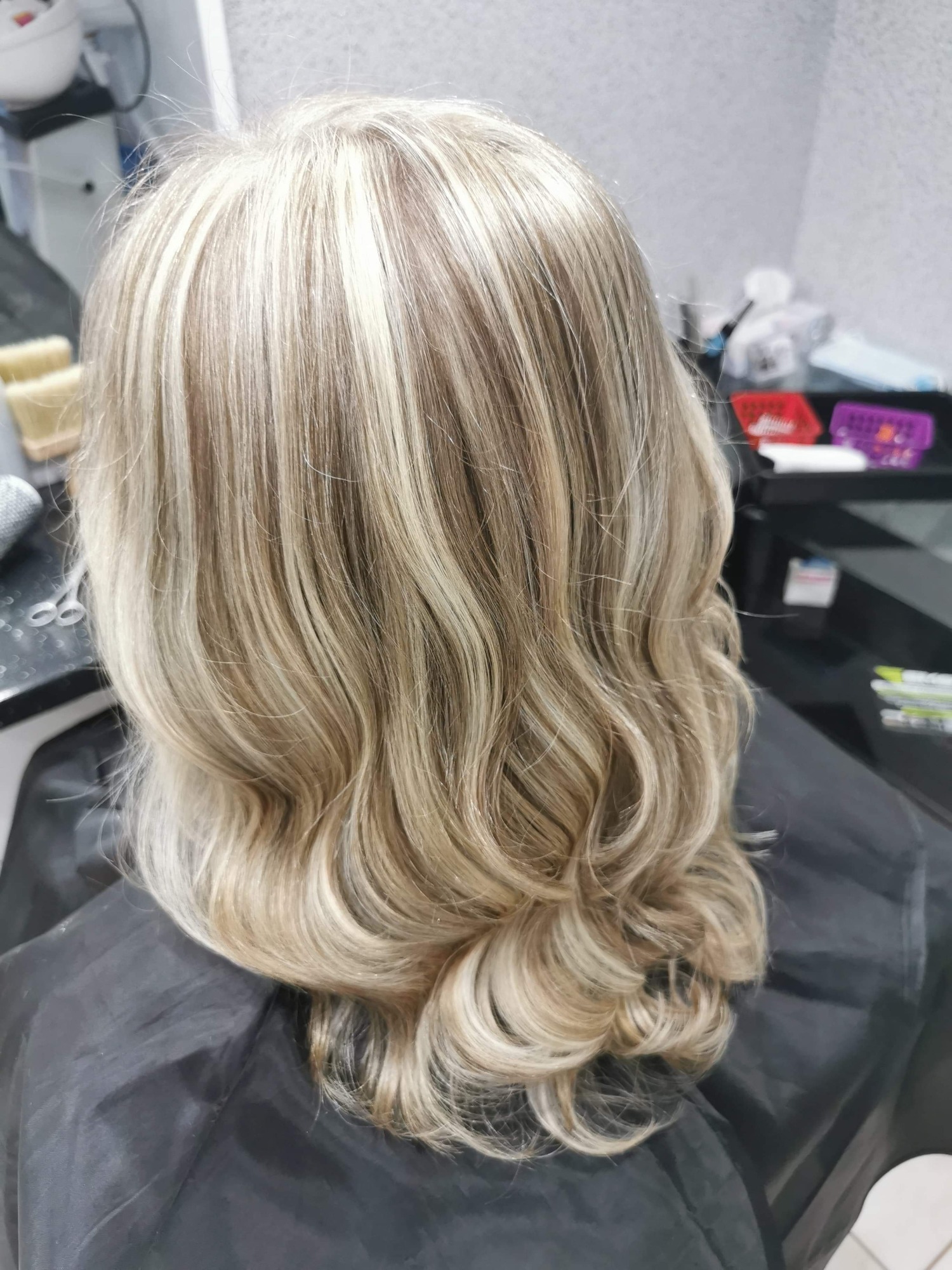 Blond włosy z pasemkami, delikatne fale, widok z tyłu na pelerynę fryzjerską w salonie