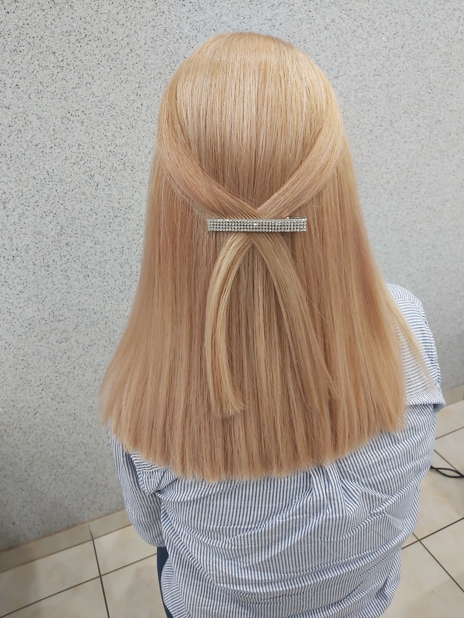 Blond włosy upięte spinką z cyrkoniami, widok z tyłu na tle szarej ściany i kafelkowej podłogi, kobieta w bluzce w niebieskie paski.