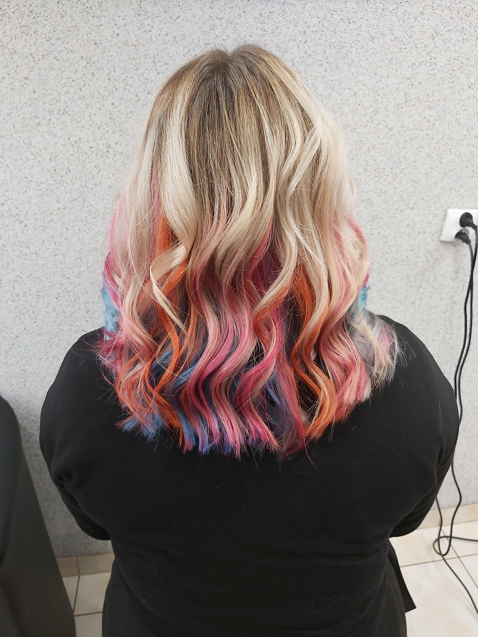 Tył głowy kobiety z blond, falowanymi włosami z kolorowymi pasemkami w odcieniach różu, pomarańczy i błękitu, widoczne gniazdko elektryczne na ścianie.