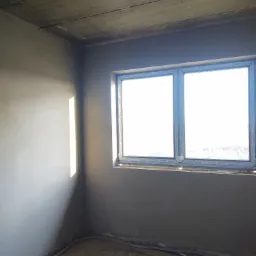 Pokój po nałożeniu tynków maszynowych, widoczne świeżo otynkowane ściany, surowy betonowy sufit i okno z plastikową ramą Gealan, na podłodze widoczne kable.