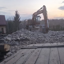 WBT DYNAMIK SŁAWOMIR SZAFRANIEC - Duża koparka kołowa Caterpillar stoi na rozległym gruzowisku po rozbiórce budynku, z drewnianymi deskami na pierwszym planie, o zmierzchu w Bochni.
