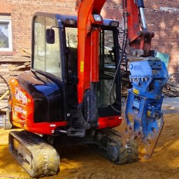 WBT DYNAMIK SŁAWOMIR SZAFRANIEC - Czerwono-czarna minikoparka Kubota KX030-4 z niebieskim osprzętem hydraulicznym do rozbiórki, na placu z gruzem ceglanym, w tle ceglany mur budynku oświetlony słońcem.