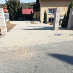 Nowo utwardzony podjazd do domu z betonowych płyt, widoczne słupki wejściowe z szarego kamienia i częściowo zagospodarowany teren wokół, dom w tle.