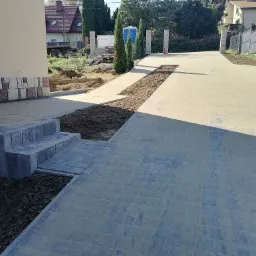 Ułożona kostka brukowa na podjeździe, schody wejściowe z kostki, widoczne przygotowane rabaty ziemne po bokach, w tle budynki mieszkalne i przenośna toaleta.