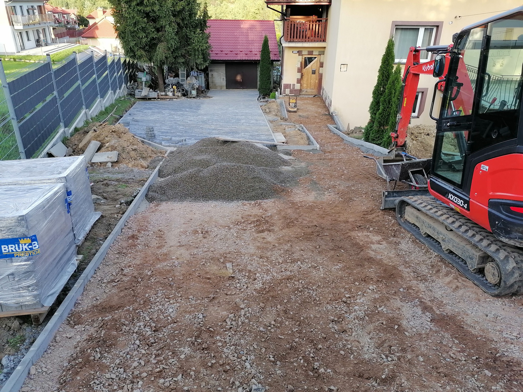 Prace przygotowawcze do ułożenia kostki brukowej w Bochni, widoczny teren wyrównany, paleta z kostką Bruk-Bet Prestige, mini koparka Kubota KX030-4 oraz częściowo ułożona nawierzchnia z kostki.