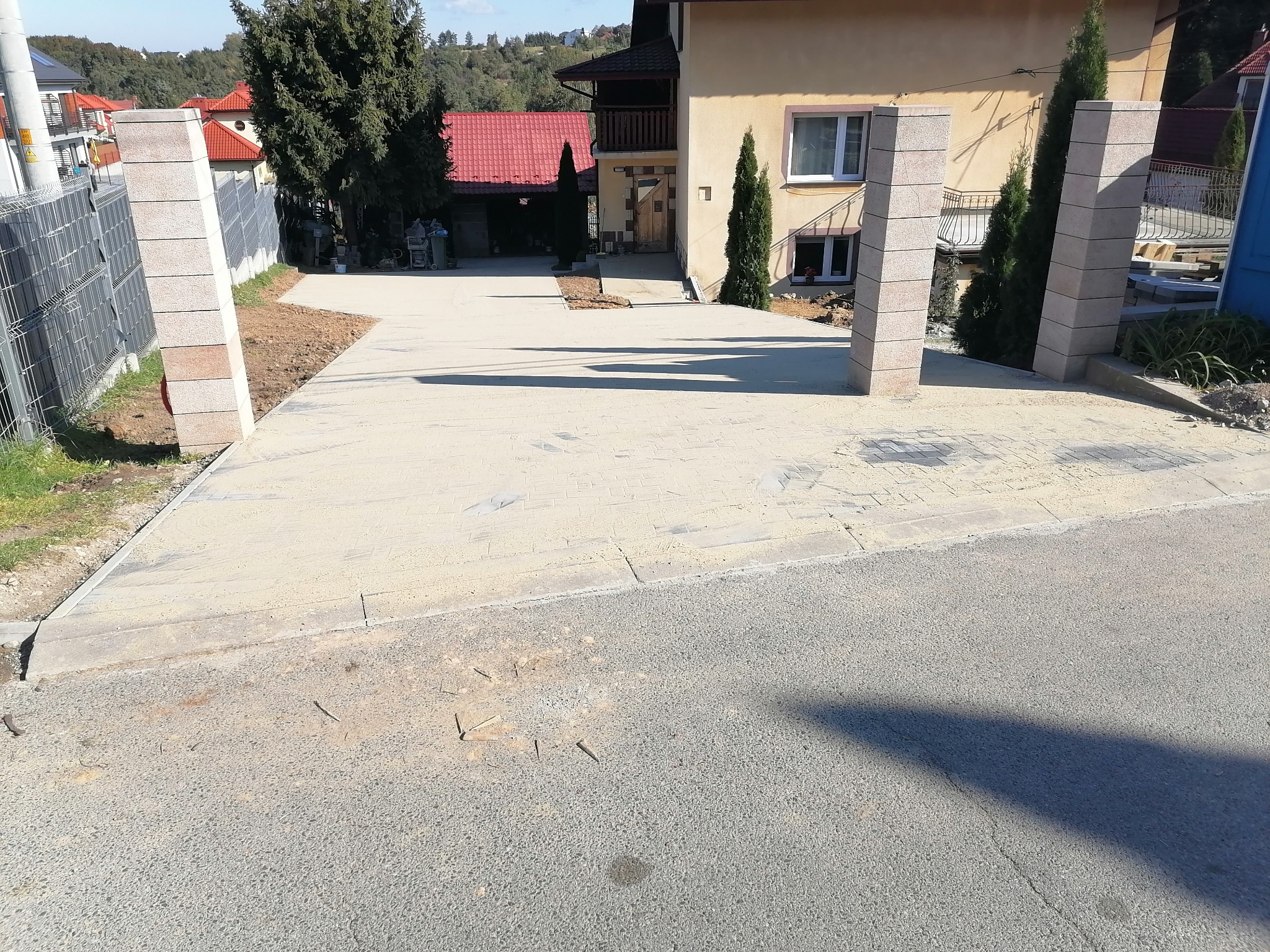 Nowo utwardzony podjazd do domu z betonowych płyt, widoczne słupki wejściowe z szarego kamienia i częściowo zagospodarowany teren wokół, dom w tle.