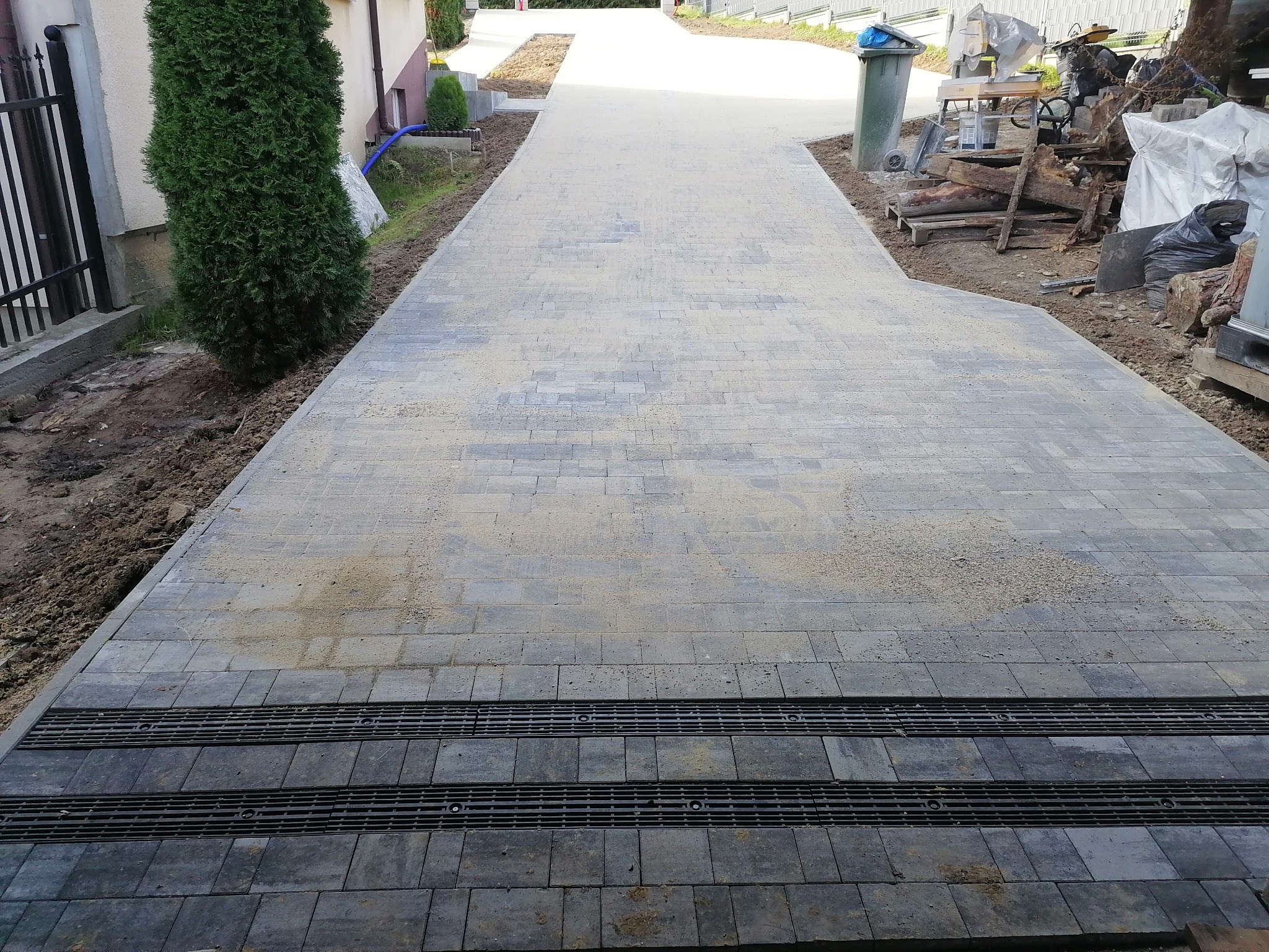 Szeroki podjazd z szarej kostki brukowej z liniowym odwodnieniem, posypany piaskiem, widok z dołu w kierunku ulicy, obok podjazdu rosną tuje.