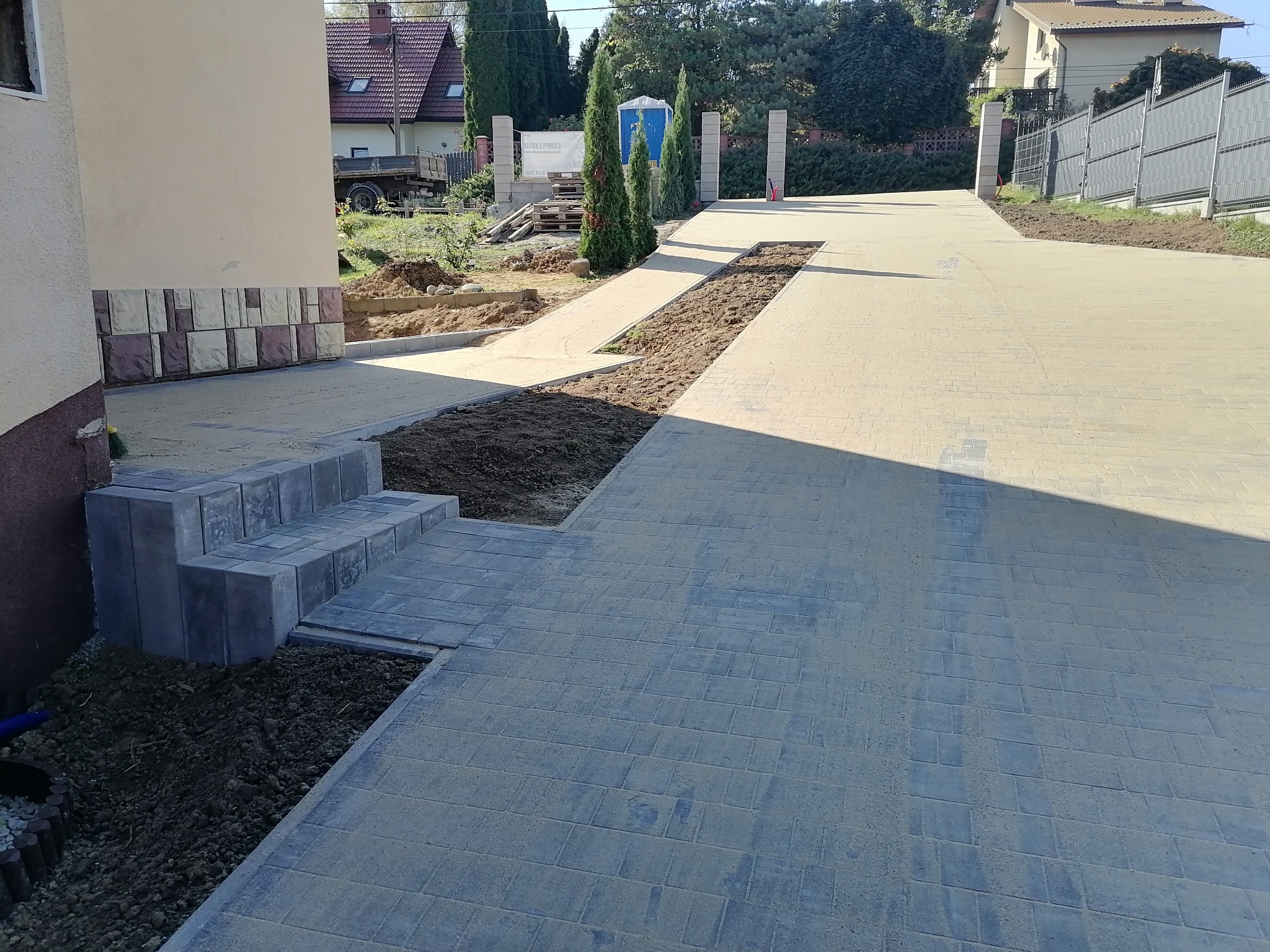 Ułożona kostka brukowa na podjeździe, schody wejściowe z kostki, widoczne przygotowane rabaty ziemne po bokach, w tle budynki mieszkalne i przenośna toaleta.