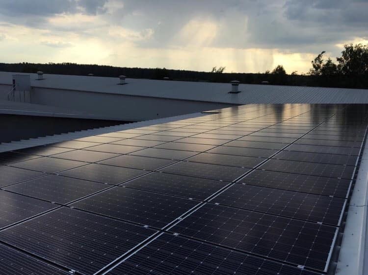 Panele słoneczne na dachu budynku przemysłowego, widoczne kominki wentylacyjne i zbliżająca się burza w tle.