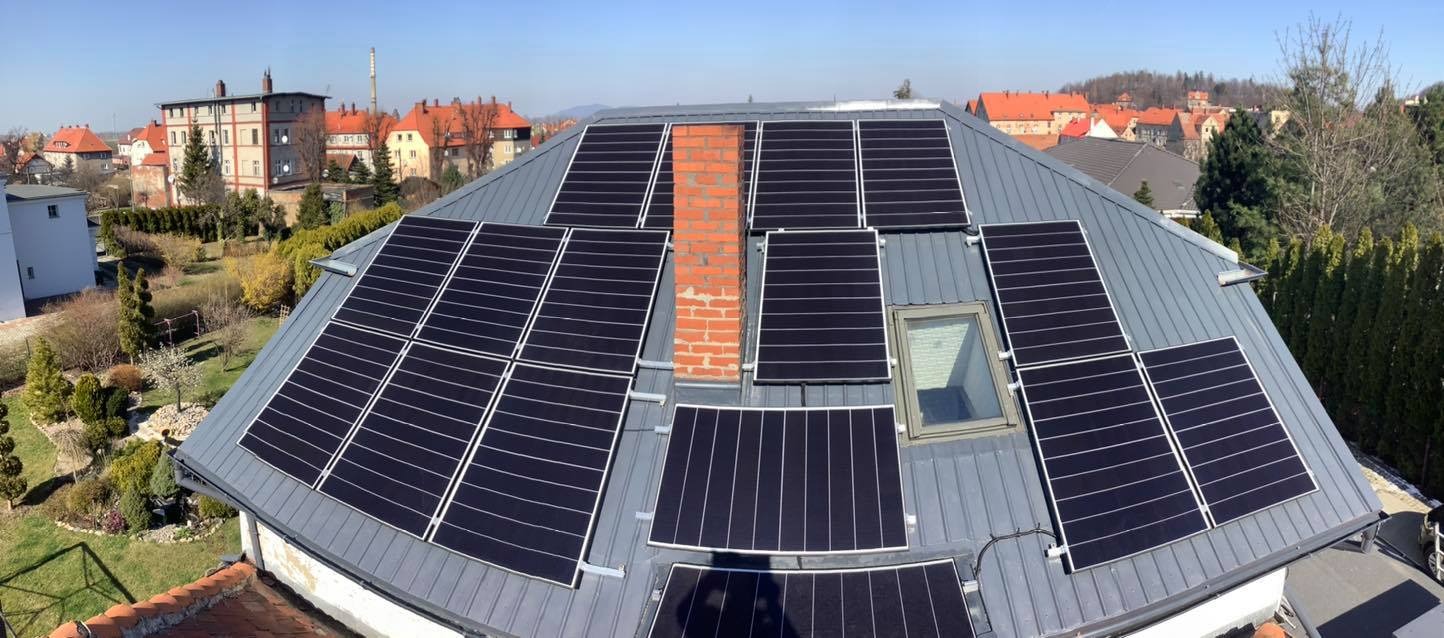 Panele słoneczne na szarym blaszanym dachu domu jednorodzinnego, z kominem i oknem dachowym, w tle zabudowa miejska.