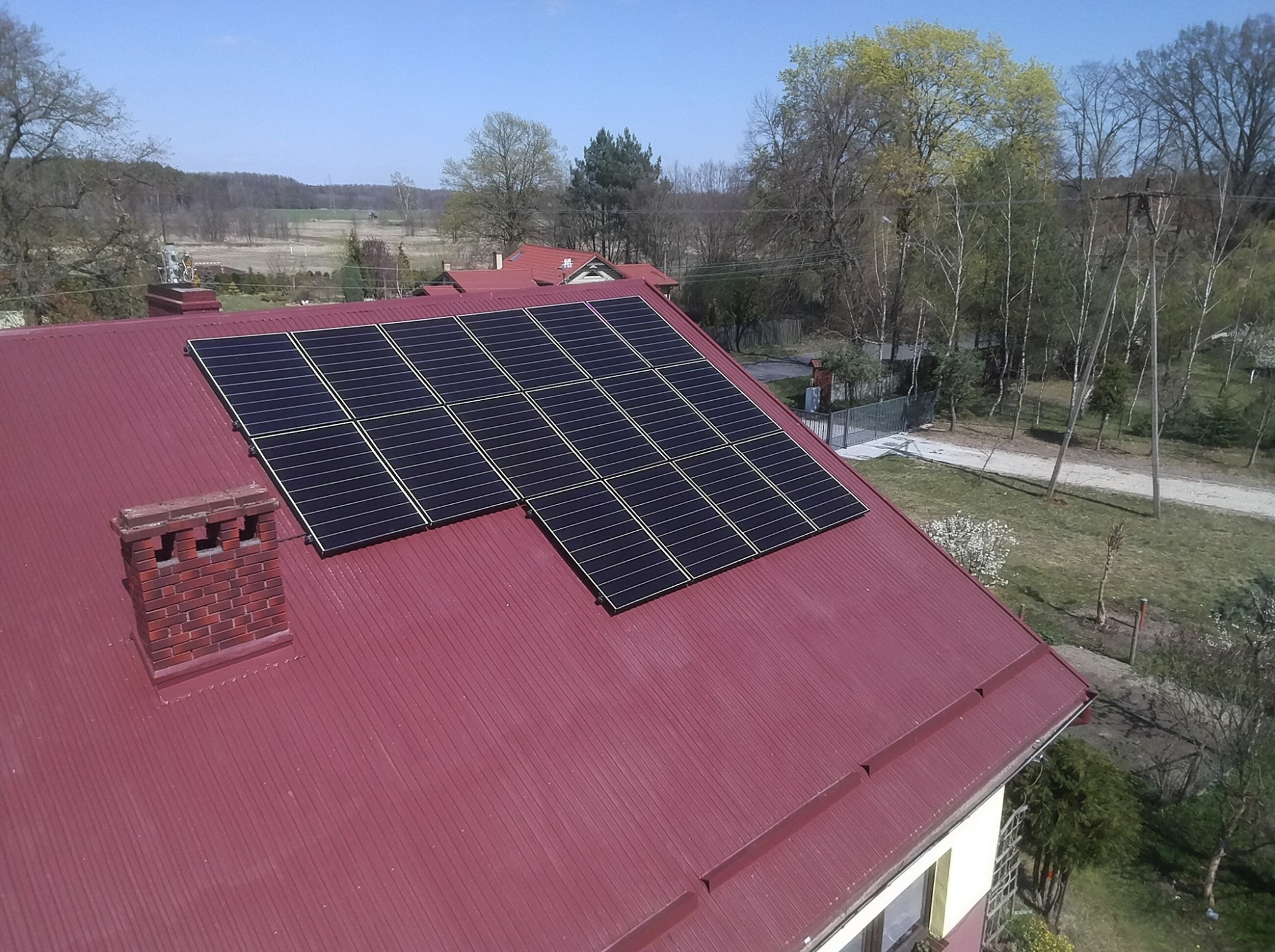 Panele słoneczne na dachu domu z ceglanym kominem, widok z góry, w tle drzewa i zabudowa wiejska.