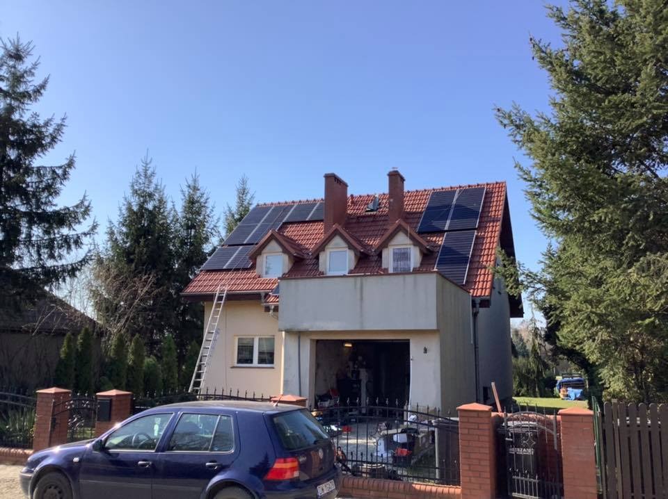 Dom jednorodzinny z czerwoną dachówką i panelami słonecznymi na dachu, drabina oparta o ścianę, niebieski samochód zaparkowany przed posesją.