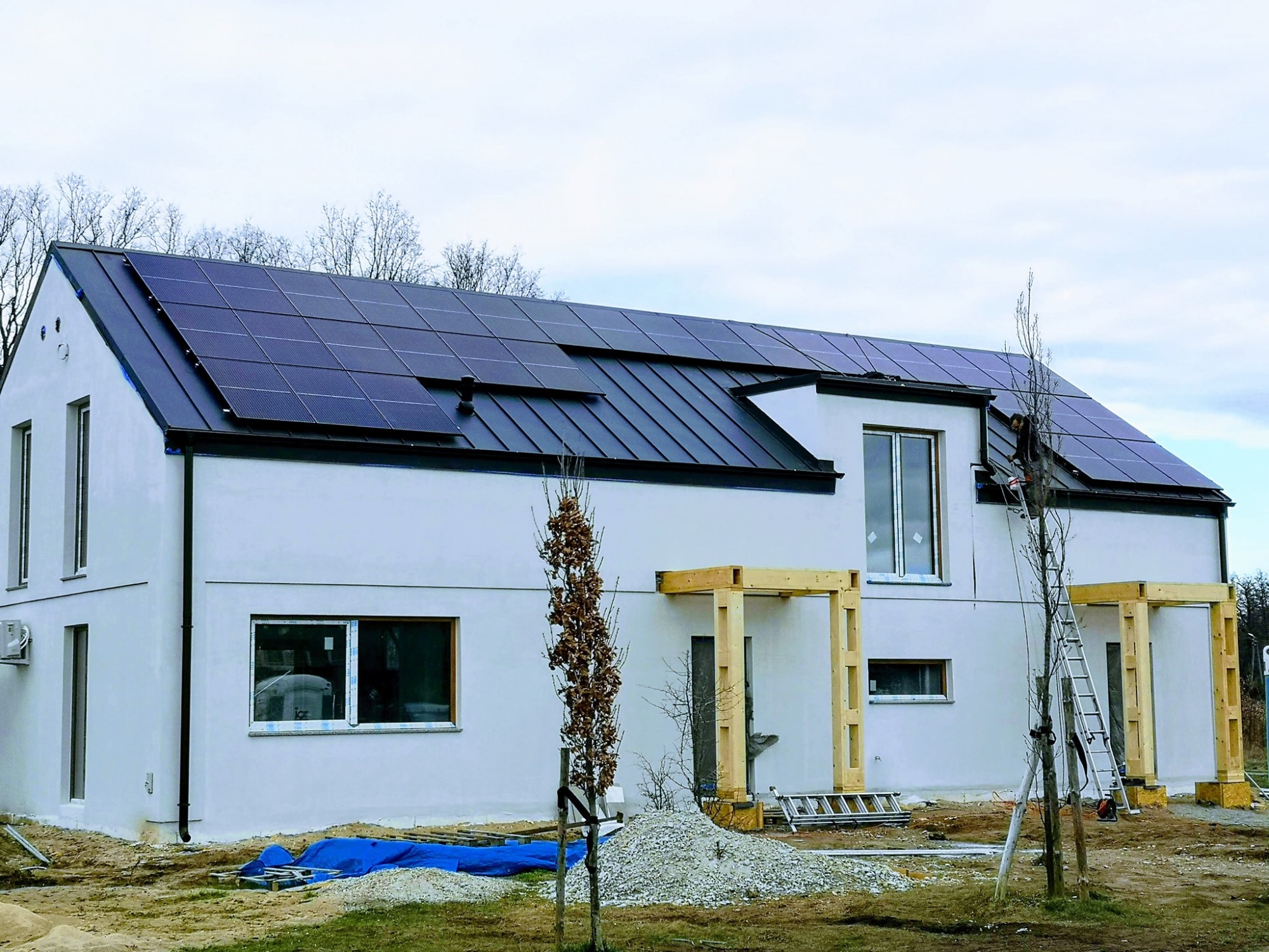 Nowoczesny dom jednorodzinny z panelami słonecznymi na dachu, częściowo zamontowanymi, z pracownikiem na drabinie, drewniane konstrukcje ganku w trakcie budowy, plac budowy.