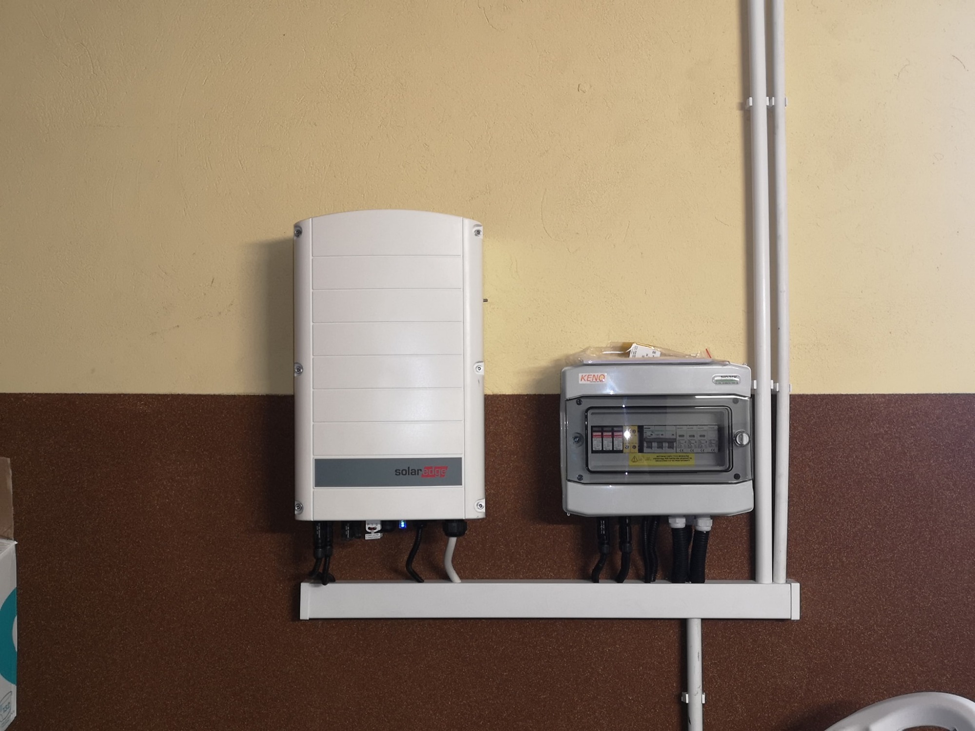 Inwerter fotowoltaiczny SolarEdge i skrzynka bezpiecznikowa KENO zamontowane na dwukolorowej ścianie z brązową boazerią i beżową górą, z widocznymi kablami i rurami instalacyjnymi.