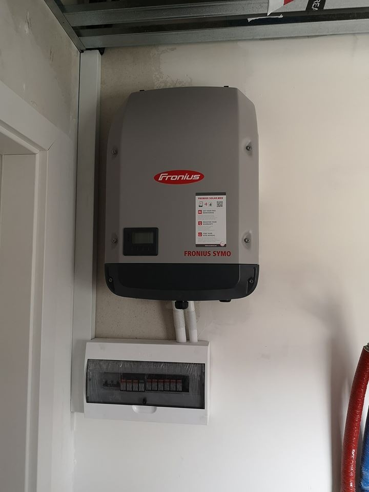 Szary falownik Fronius Symo zamontowany na białej ścianie obok skrzynki elektrycznej, widoczne rurki instalacyjne i fragment czerwonego węża.