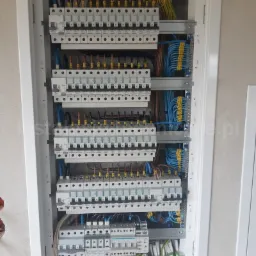 Rozdzielnica elektryczna w białej obudowie, widoczne liczne bezpieczniki, przewody w kolorach żółtym, niebieskim i brązowym, zamontowana na surowej ścianie w trakcie prac wykończeniowych.