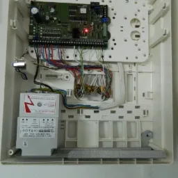 Otwarta skrzynka centrali alarmowej z widoczną płytą główną, transformatorem i podłączonymi przewodami, zamontowana na ścianie.