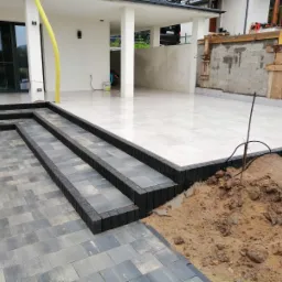 Ułożone schody z kostki brukowej prowadzące do tarasu z jasnych płytek, widoczny fragment domu z białymi ścianami i słupami, obok hałda ziemi.