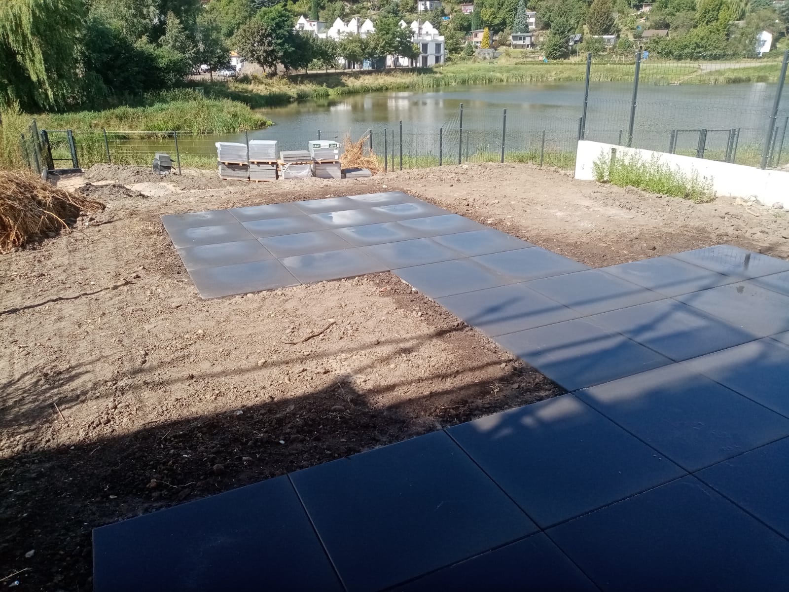 Fragment układanej nawierzchni z ciemnoszarych płyt betonowych na gruncie, w tle palety z kostką i zbiornik wodny.