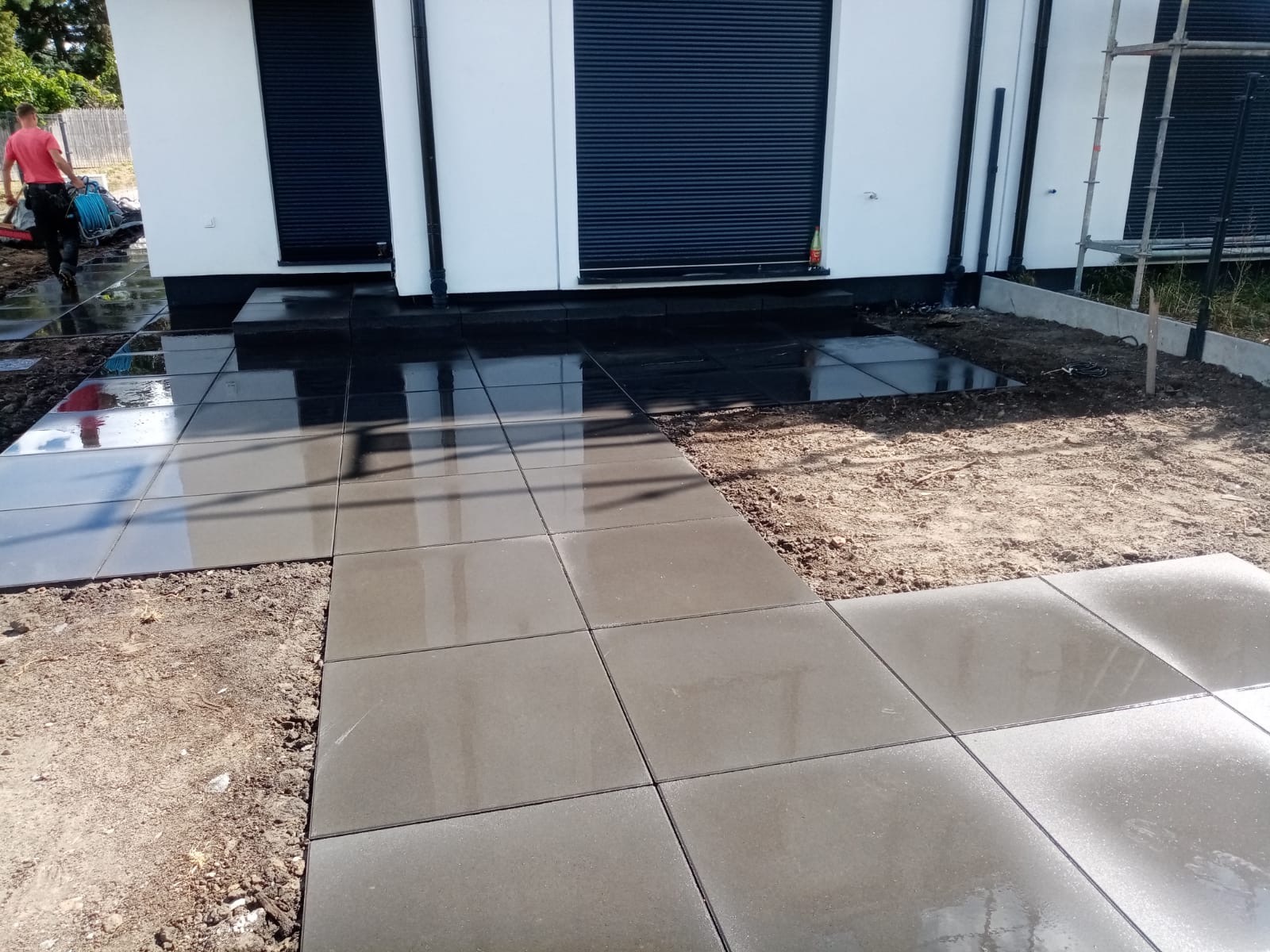 Układanie szarych, kwadratowych płyt chodnikowych na posesji, widoczny fragment budynku z czarnymi roletami i pracownik w oddali.