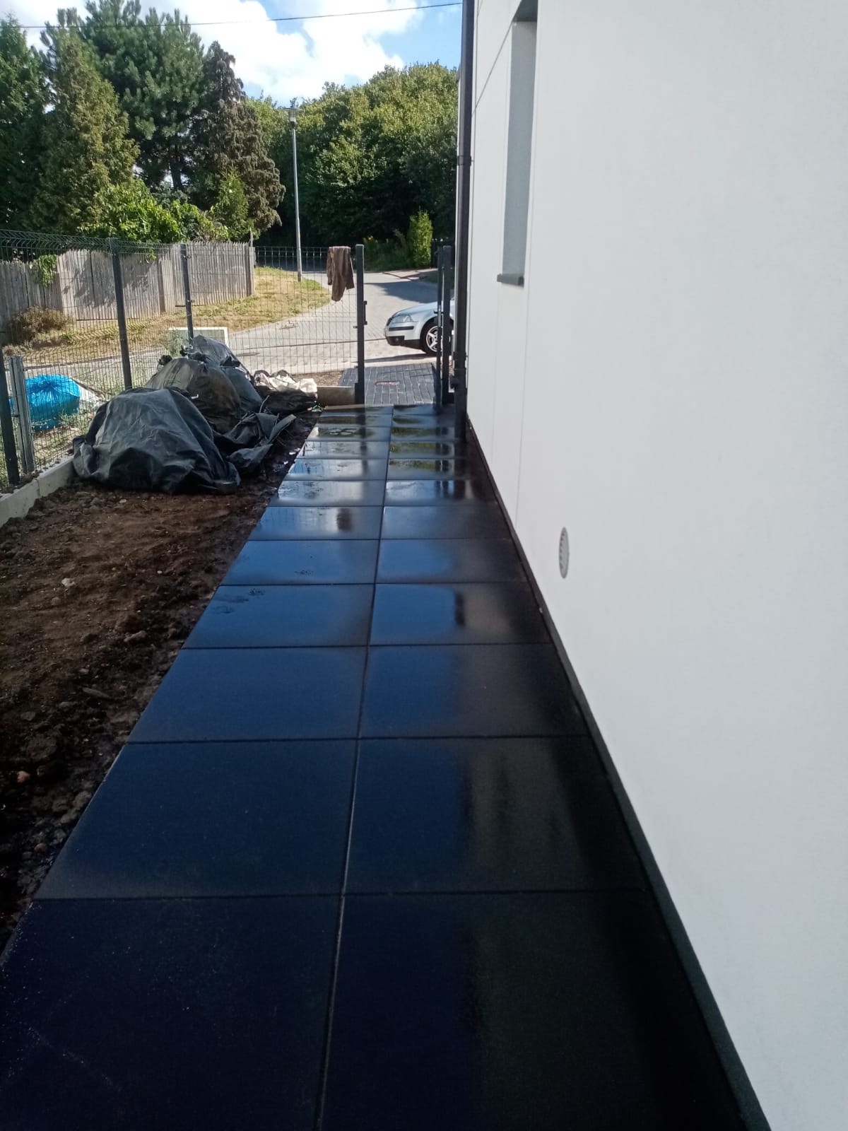 Świeżo ułożony, błyszczący chodnik z ciemnych płyt przy białej ścianie domu. Widoczne worki z materiałem budowlanym przy ogrodzeniu i fragment zaparkowanego samochodu w tle.