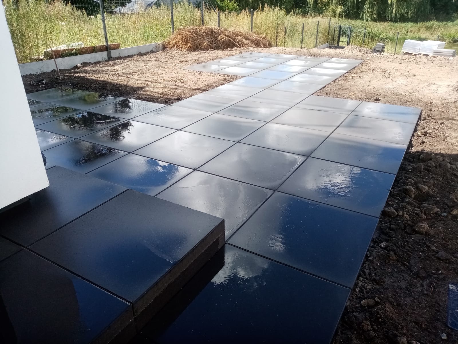 Świeżo ułożona, mokra nawierzchnia z ciemnych, kwadratowych płyt betonowych, z widocznym odbiciem nieba, obok fragmentu białej ściany budynku i przygotowanym podłożem pod dalszą część tarasu.