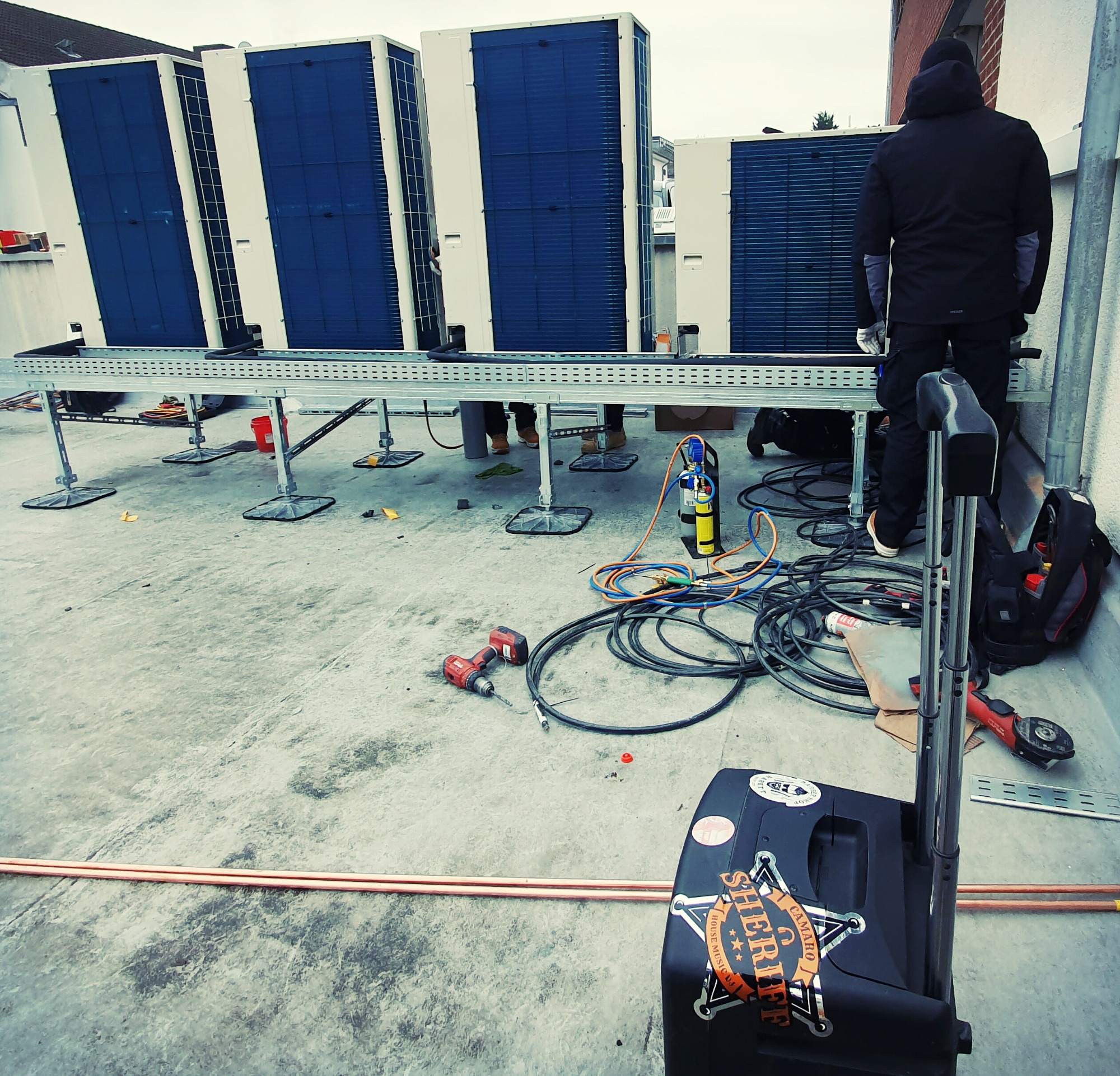 Instalacja czterech klimatyzatorów na dachu budynku, widoczny monter w czarnej kurtce, narzędzia pracy, przewody i butla z gazem.