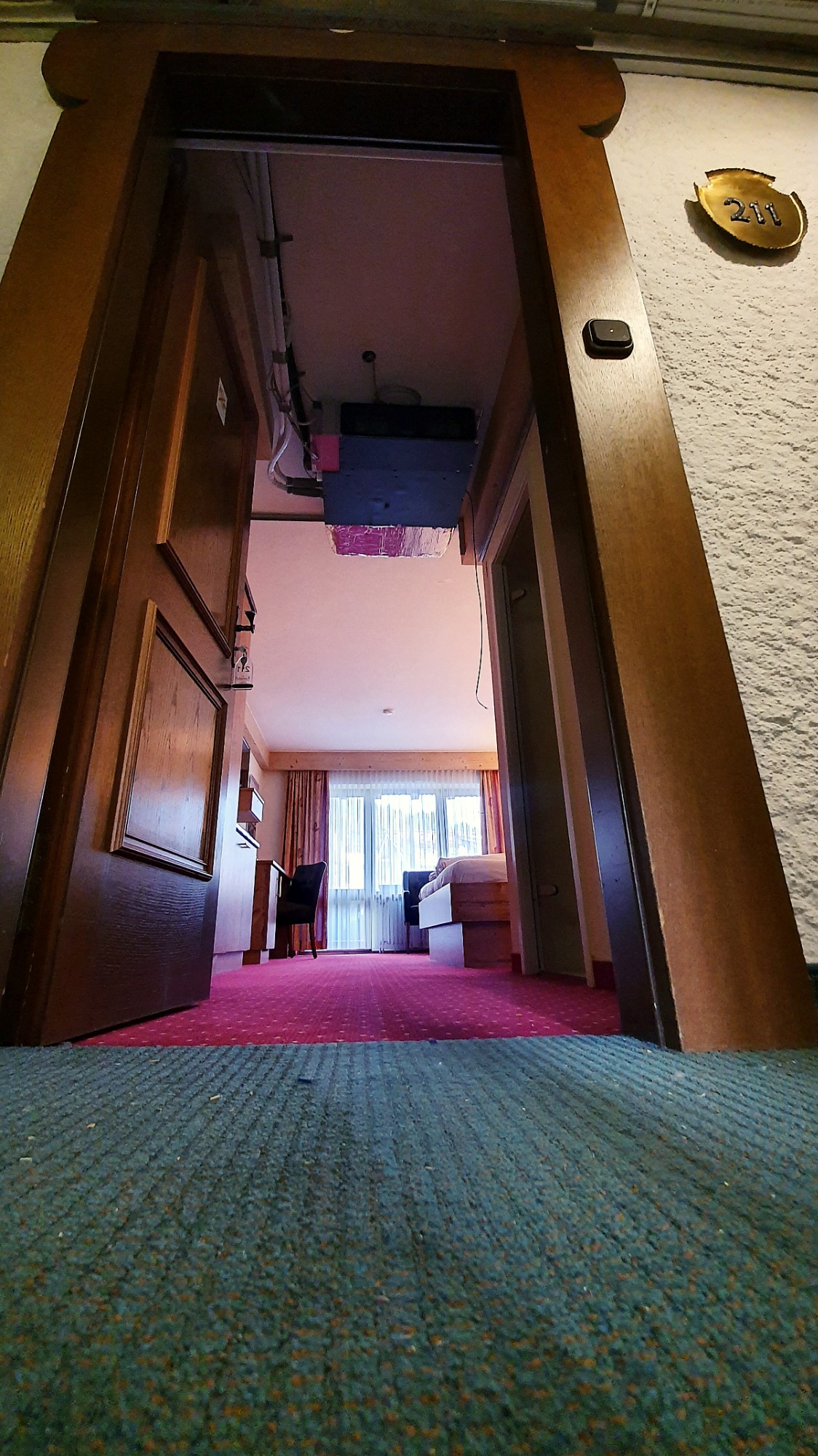 Widok z dołu na otwarte drzwi pokoju hotelowego z widocznym klimatyzatorem pod sufitem, biurkiem i łóżkiem w oddali.