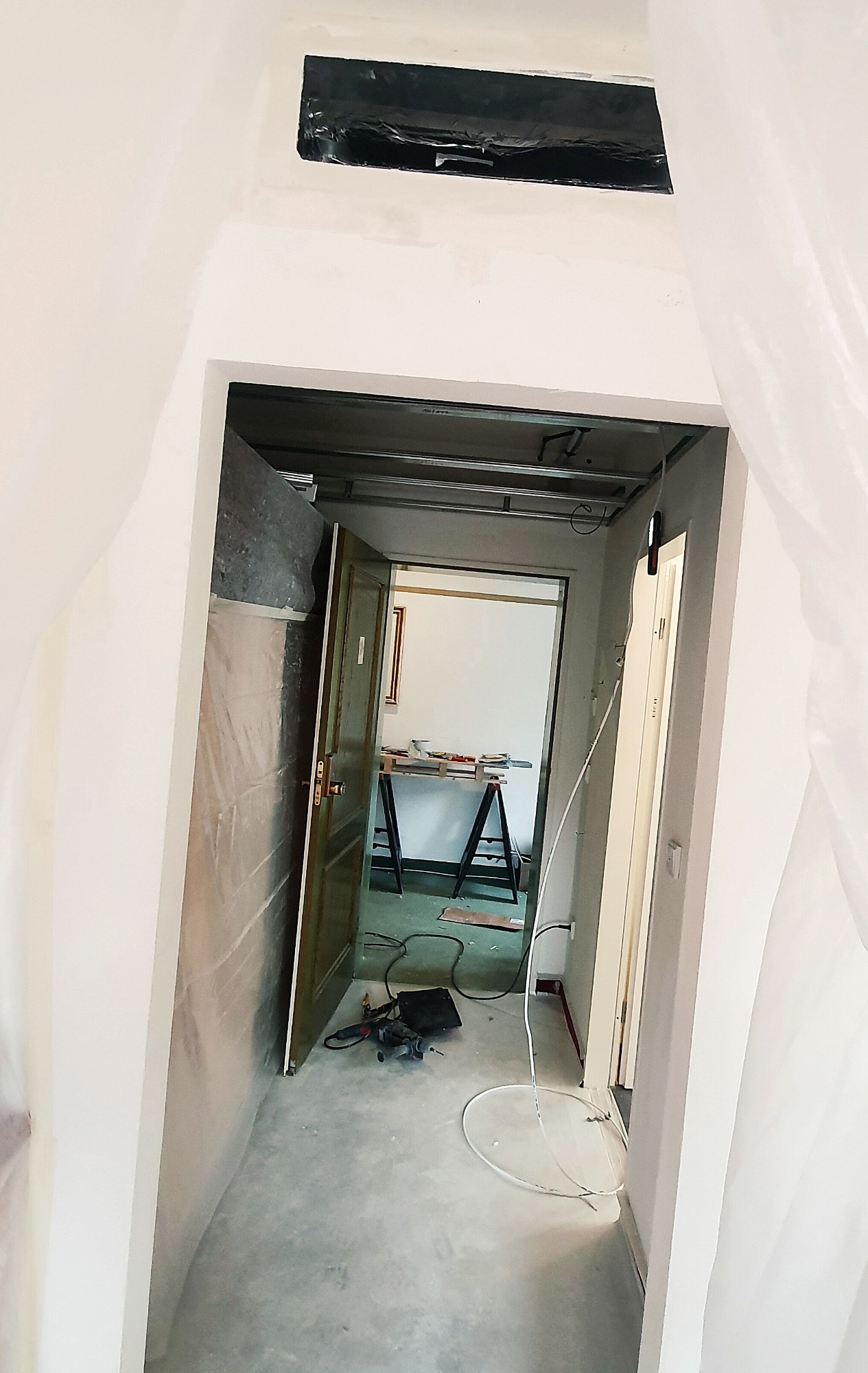 Wnętrze w trakcie remontu, widoczny otwór wentylacyjny zabezpieczony folią, podwieszany sufit w trakcie montażu, narzędzia na podłodze i drabina w oddali.