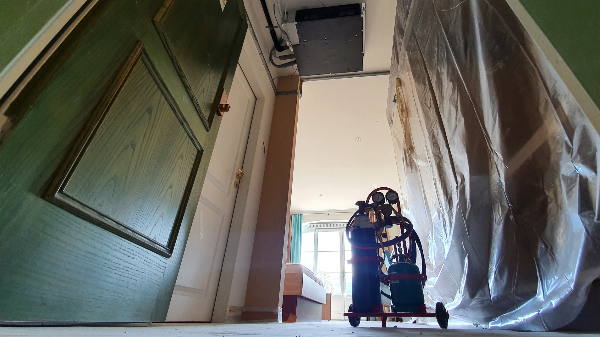 Instalacja klimatyzacji w domu: widok z dołu na butle z gazem, zabezpieczoną folią przestrzeń i zamontowaną jednostkę klimatyzacyjną pod sufitem, z widocznym fragmentem sypialni w tle.