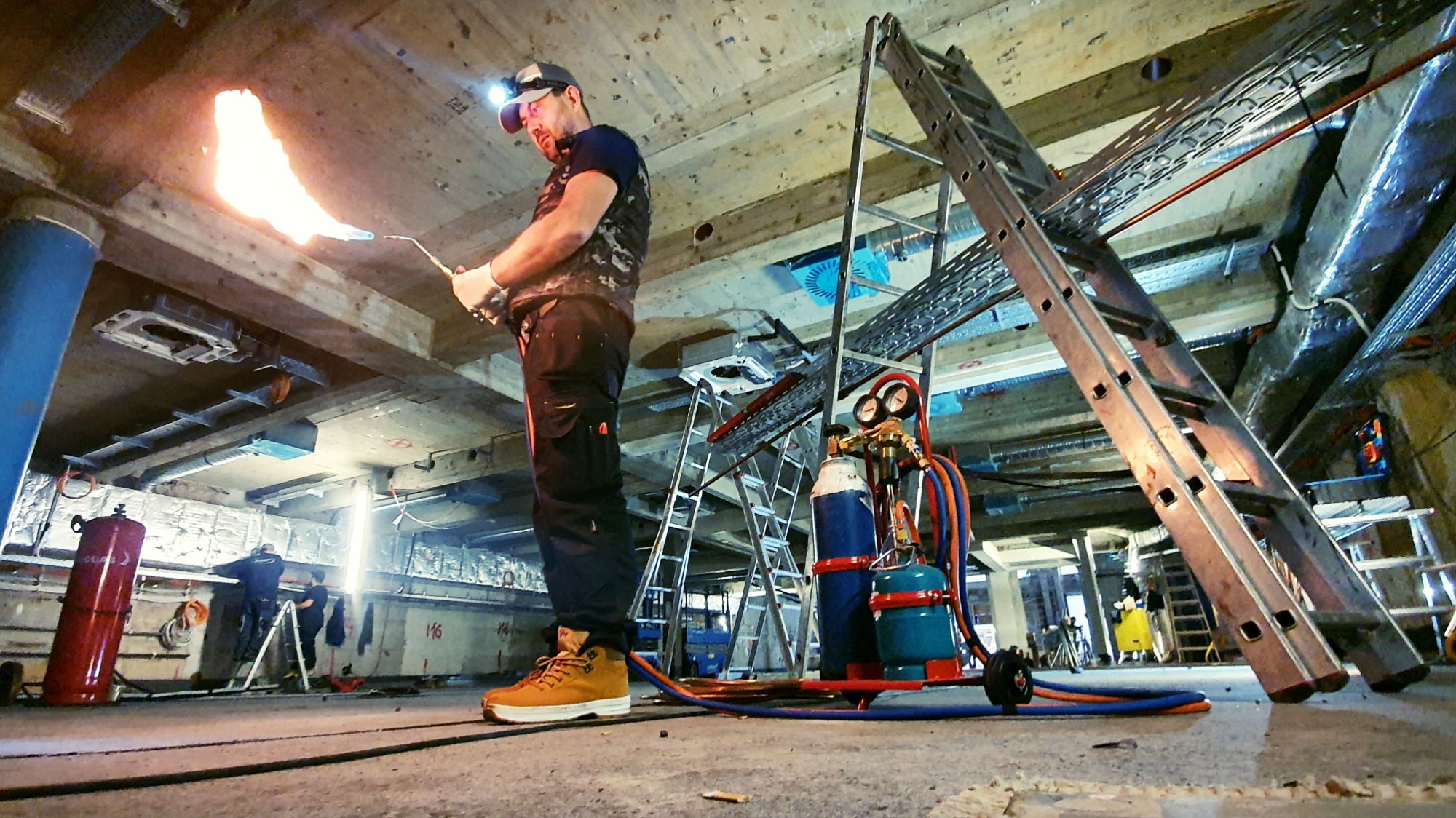 Pracownik w kasku z czołówką lutuje palnikiem gazowym elementy pod sufitem, widoczne butle z gazem, drabiny i instalacje wentylacyjne.