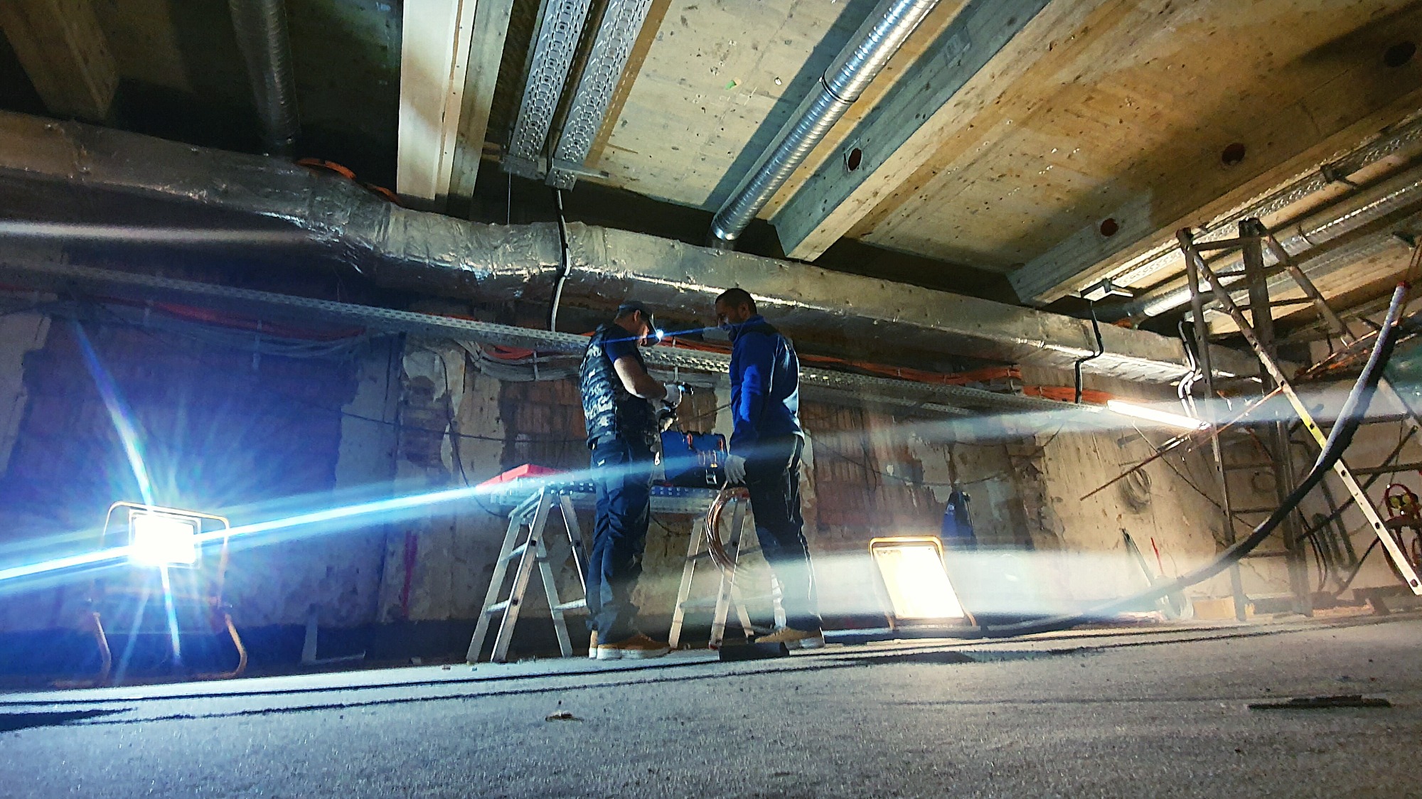 Dwóch monterów na drabinach instaluje system wentylacyjny w surowym wnętrzu, widoczne rury wentylacyjne pod drewnianym stropem, oświetlenie budowlane.