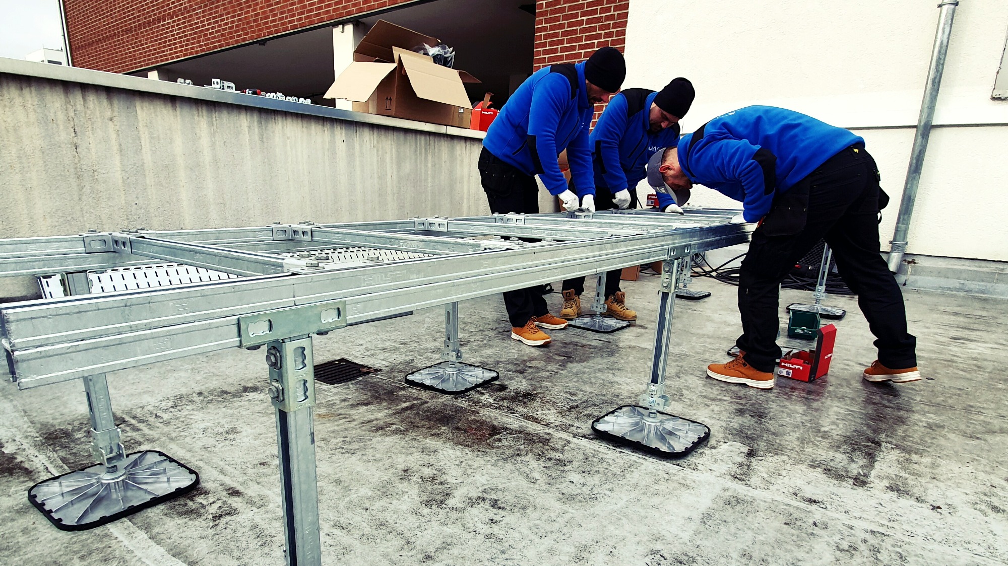 Trzech monterów w niebieskich bluzach instaluje metalową konstrukcję wsporczą na płaskim dachu, widoczna skrzynka narzędziowa Hilti.