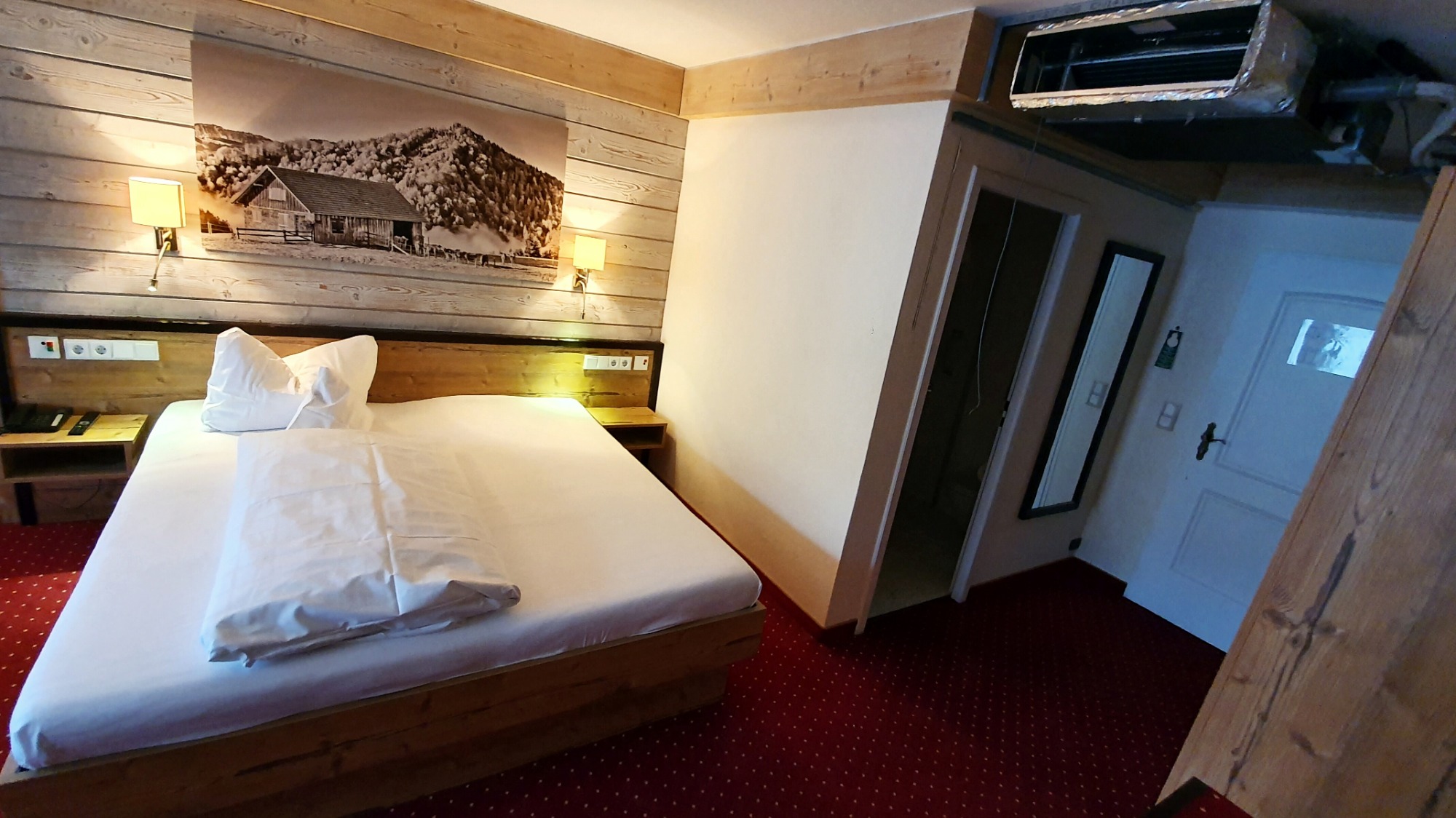 Wnętrze pokoju hotelowego z drewnianym wykończeniem, klimatyzatorem zamontowanym pod sufitem i łóżkiem z białą pościelą.