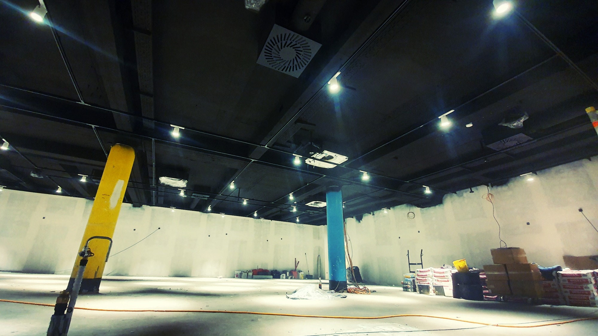 Wnętrze hali przemysłowej w trakcie montażu instalacji wentylacyjnej i klimatyzacyjnej, widoczne kanały wentylacyjne pod sufitem, kolumna pomalowana na niebiesko, żółty słup podtrzymujący...