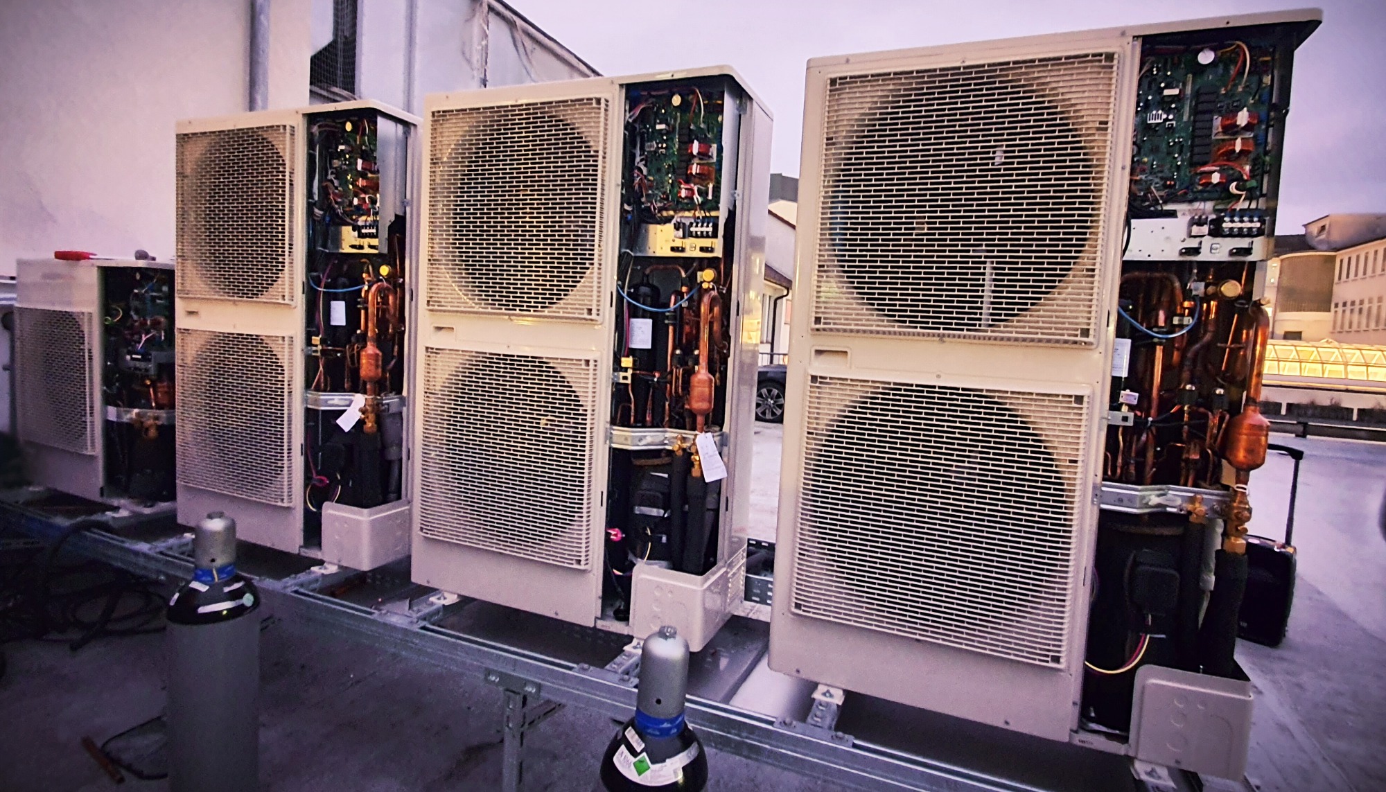 Rząd zewnętrznych jednostek klimatyzacji z otwartymi obudowami, widoczne wewnętrzne komponenty, butle z gazem chłodniczym na dachu, widok z niskiego kąta.