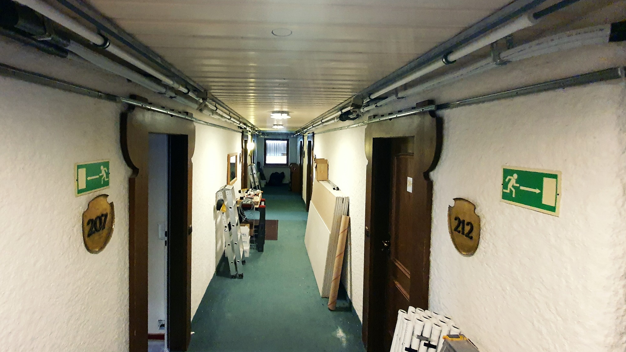 Korytarz w budynku z widocznymi rurami wentylacyjnymi pod sufitem, wyposażony w drabiny i materiały budowlane, wskazujący na trwające prace instalacyjne.