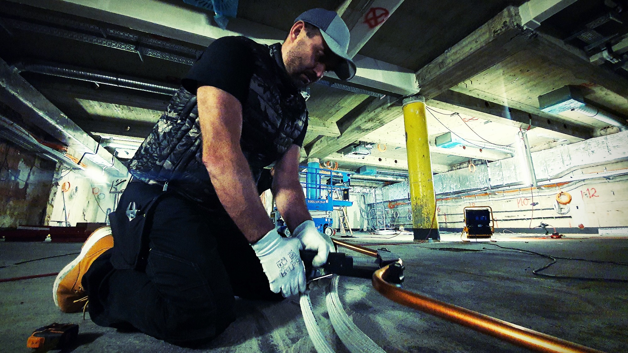 Instalator w białych rękawicach klęczy na betonowej podłodze, zaginając miedzianą rurę za pomocą specjalistycznego narzędzia; w tle widoczny podnośnik nożycowy i instalacje pod sufitem.