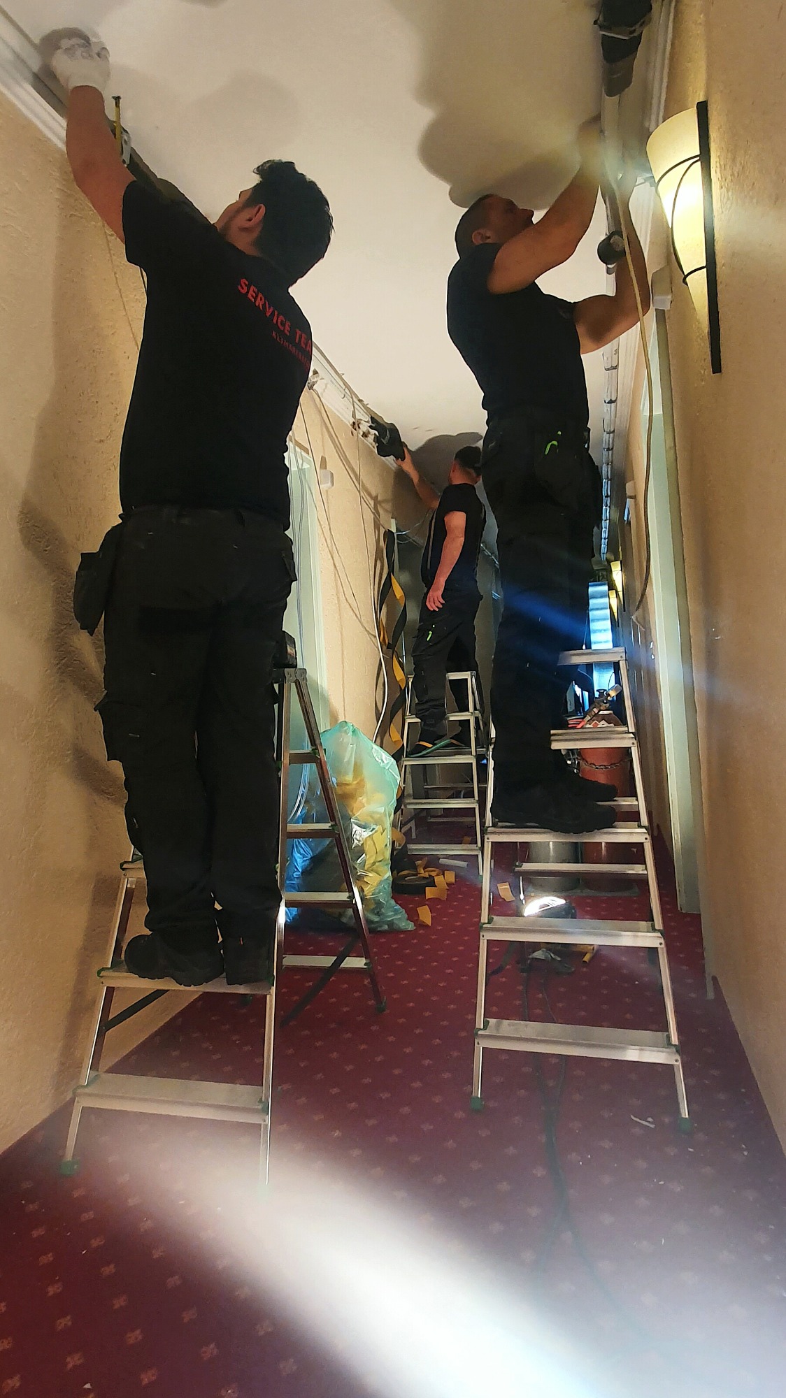 Trzech monterów w czarnych uniformach z napisem 'SERVICE TEAM' na plecach, pracujących na drabinach przy instalacji przewodów pod sufitem w korytarzu z czerwonym dywanem.