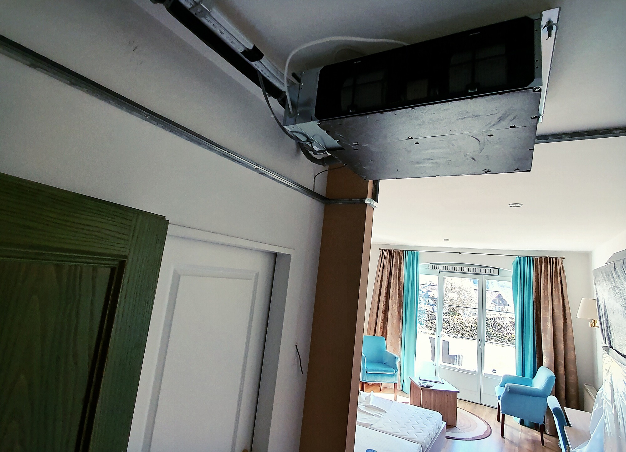 Sufitowa jednostka klimatyzacyjna w hotelowym pokoju, widoczne szyny montażowe i instalacja elektryczna, w tle okno balkonowe z widokiem na zieleń.