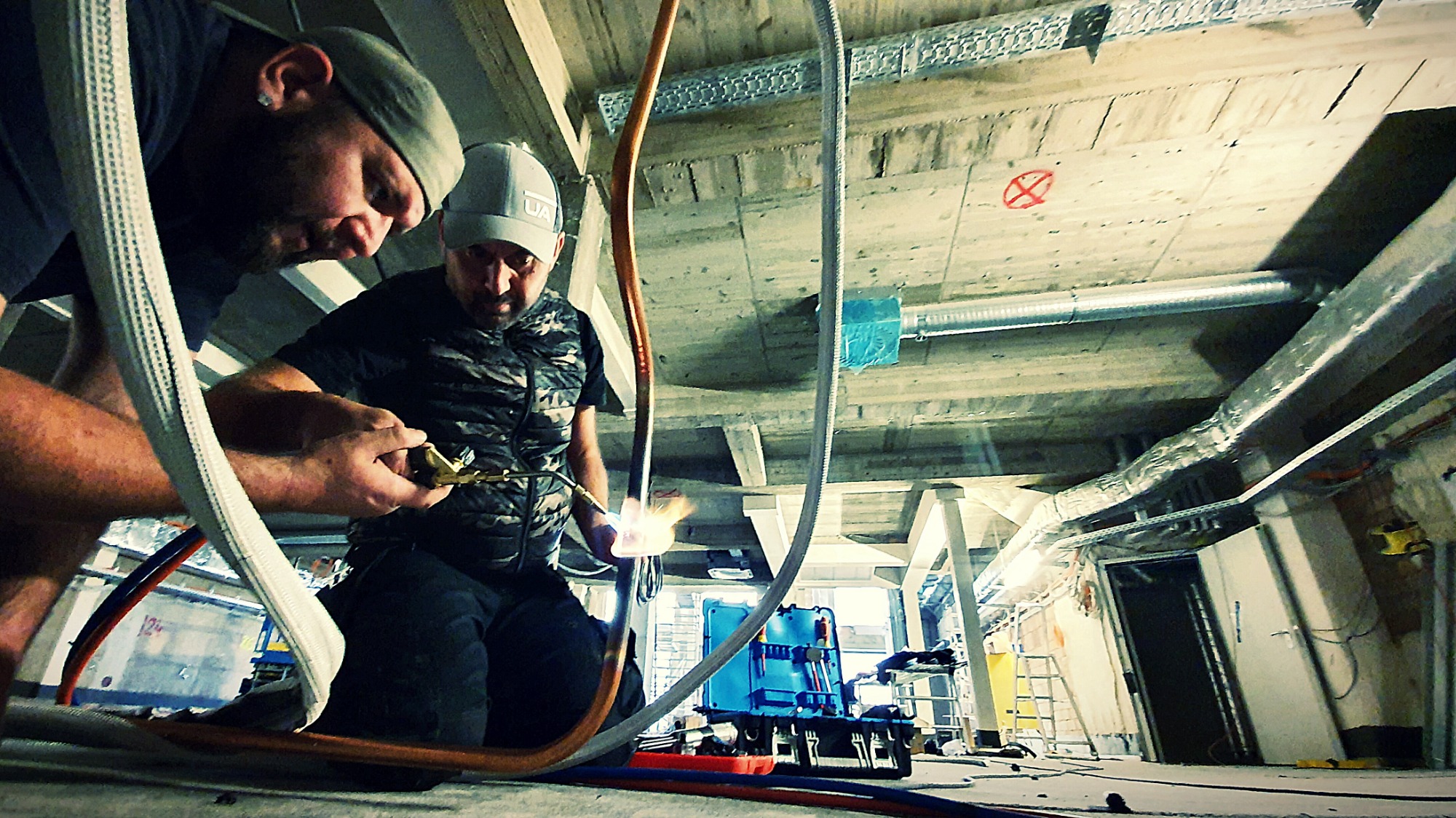 Dwóch monterów łączy rury palnikiem w surowym wnętrzu z widocznymi elementami instalacji wentylacyjnej pod sufitem.