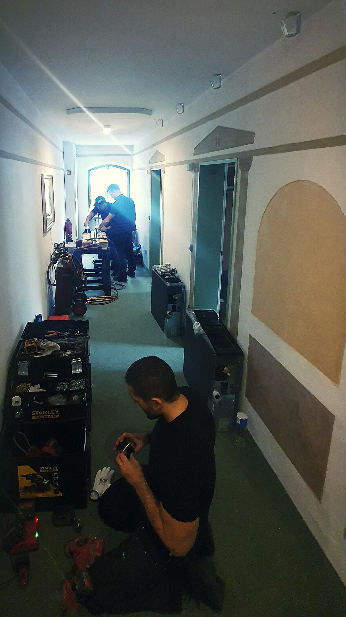 Instalator w czarnej koszulce przygotowuje się do montażu, siedząc na podłodze w korytarzu z narzędziami Stanley Fatmax obok; w tle dwóch mężczyzn pracuje przy stole warsztatowym, a wzdłuż ściany...