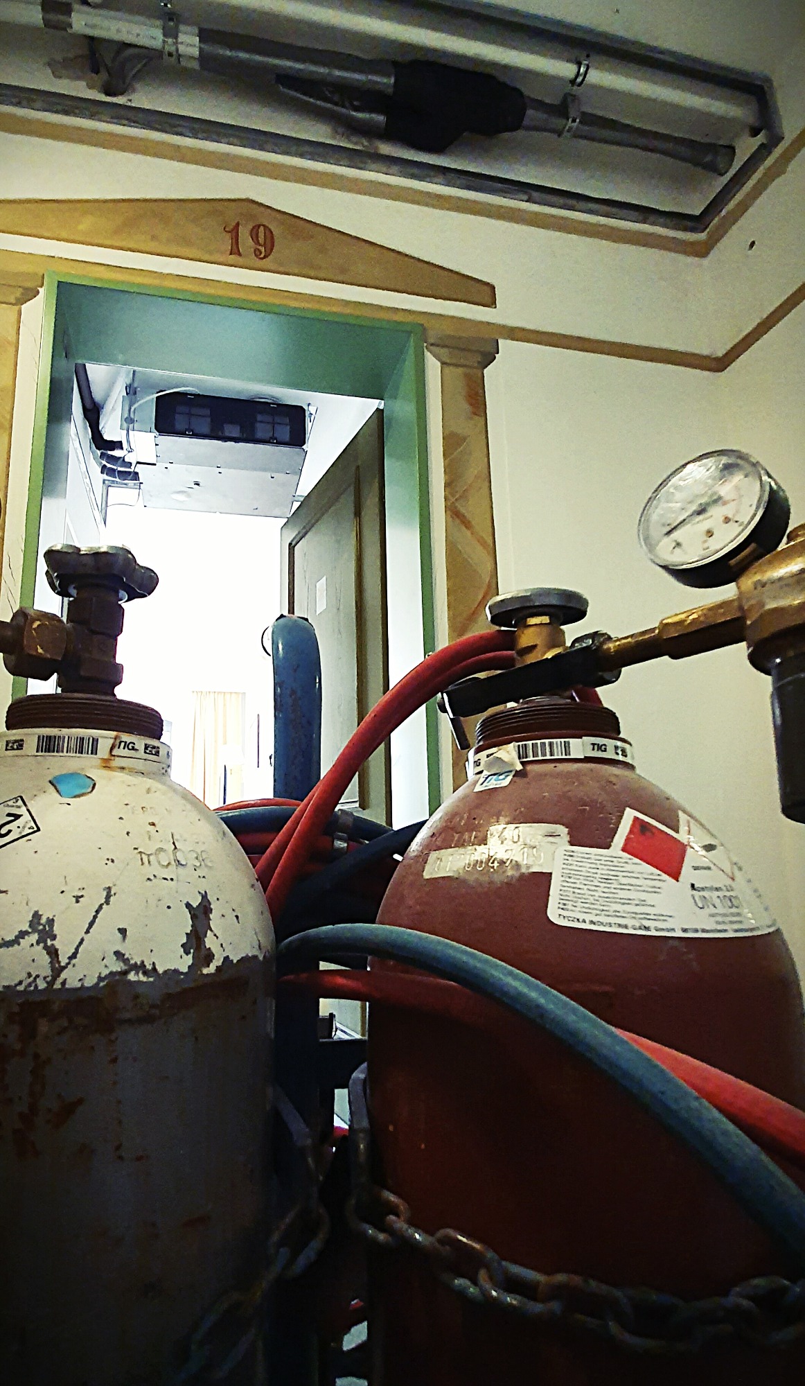 Butle z gazem technicznym z manometrem i przewodami w pomieszczeniu z zamontowaną instalacją wentylacyjną widoczną w tle.