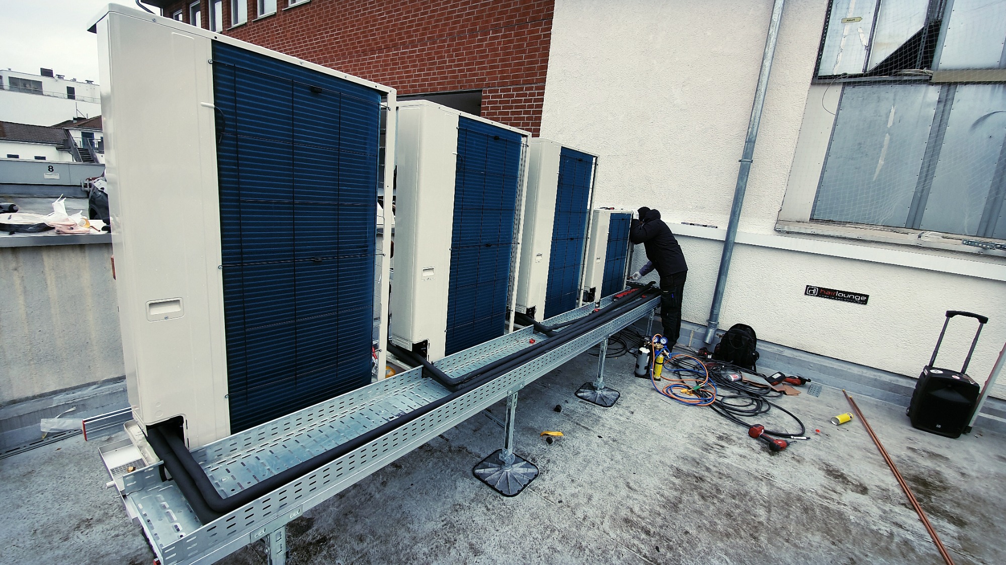 Instalacja rzędu zewnętrznych jednostek klimatyzacyjnych na dachu, widoczna osoba w trakcie pracy z narzędziami, rurociągi na metalowej konstrukcji wsporczej.