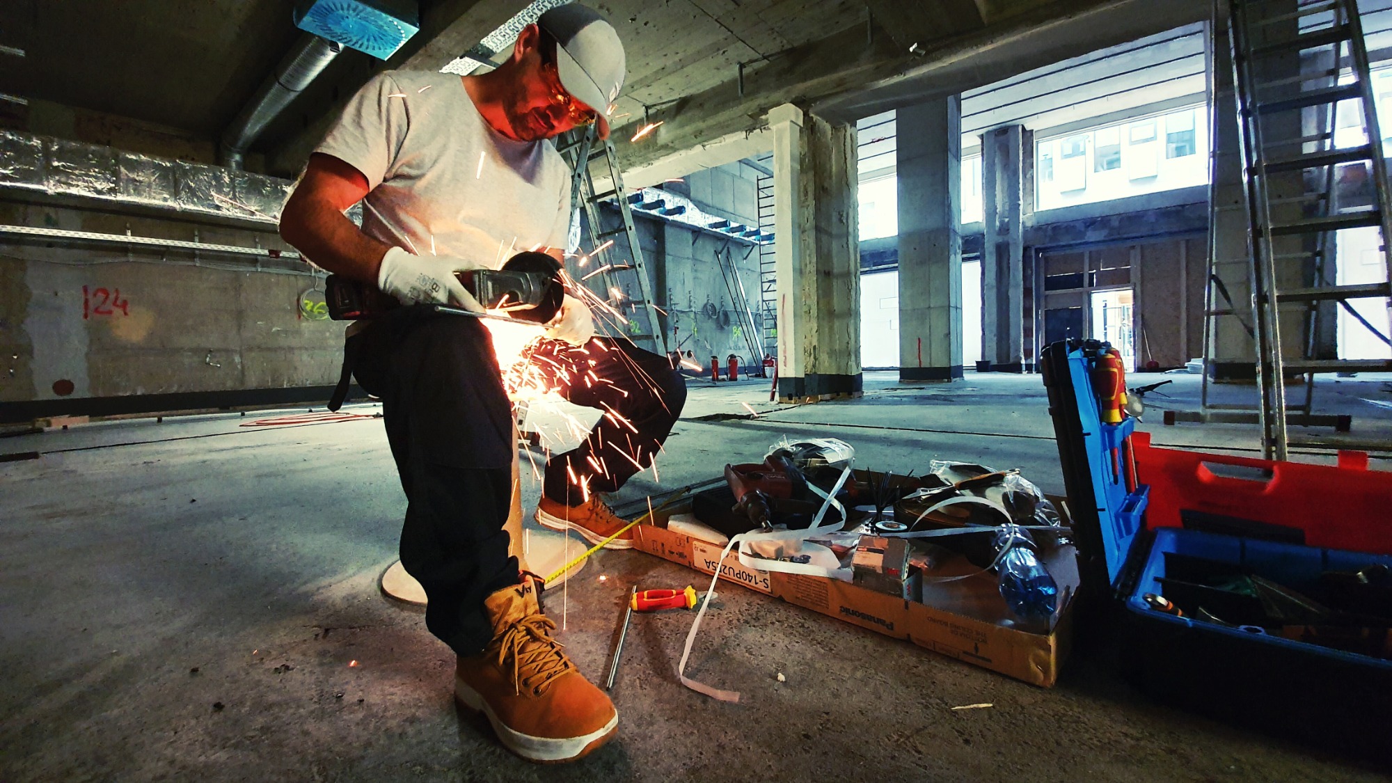 Pracownik we Wrocławiu w trakcie cięcia metalu szlifierką kątową podczas prac instalacyjnych w surowym wnętrzu budynku. W tle widoczne kolumny konstrukcyjne i drabina.