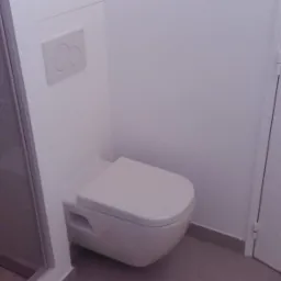 Nowoczesna toaleta podwieszana z przyciskiem spłukującym i kabiną prysznicową w tle, zamontowana w łazience w Lutczy, po remoncie.