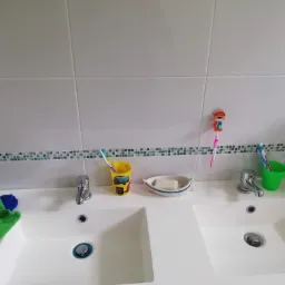 Biała łazienka z dwiema umywalkami, bateriami, kubkami z szczoteczkami do zębów i mydelniczką w kształcie łódki, wykończona białymi płytkami z zielonym paskiem mozaiki.