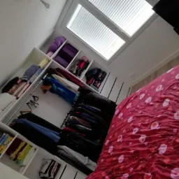 Wnętrze sypialni z białą szafą wnękową z półkami i drążkami na ubrania, widoczna pościel w czerwono-różowe wzory i okno dachowe z roletami.