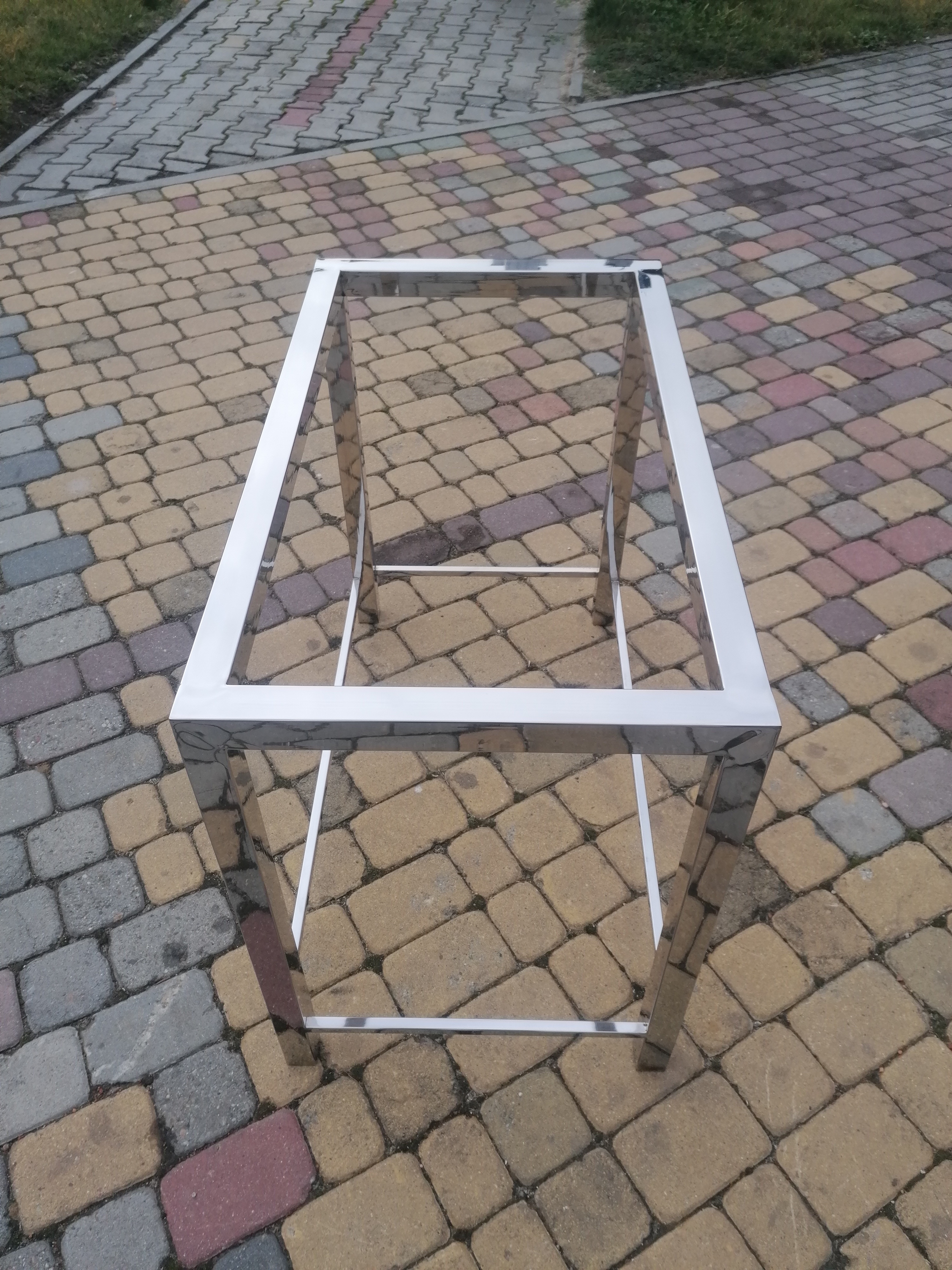 Stelaż z profili nierdzewnych, spawany, na tle kostki brukowej w odcieniach szarości, beżu i różu.