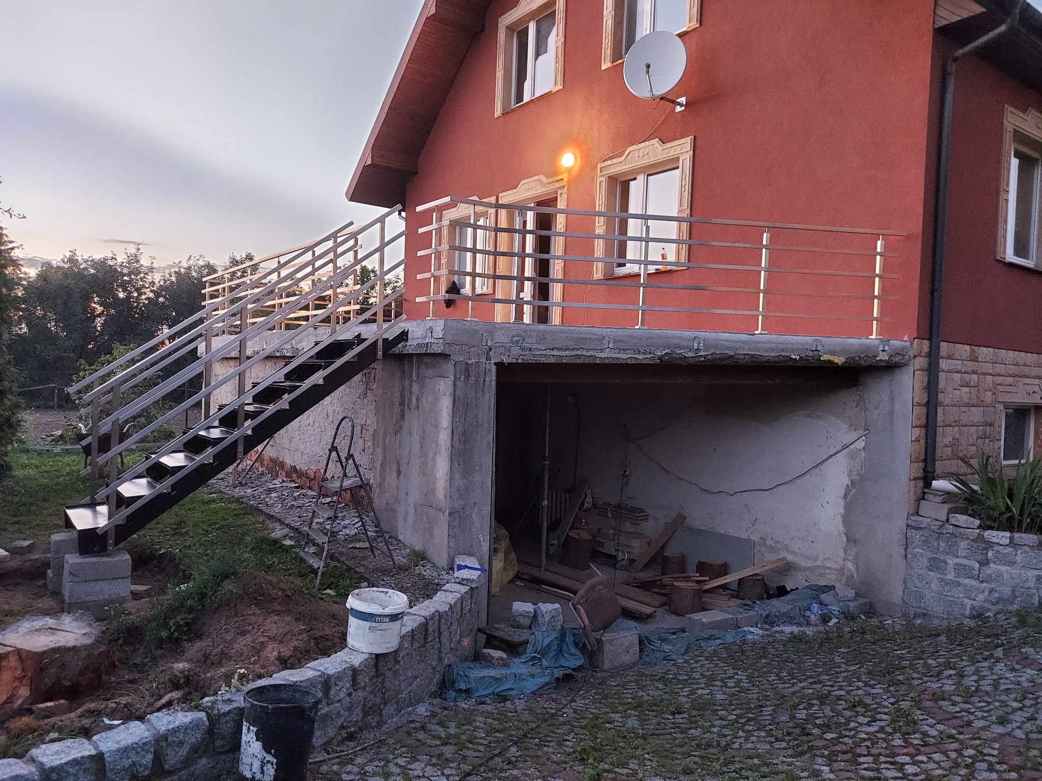 Zewnętrzne schody z metalową konstrukcją i balustradą prowadzące na balkon domu z czerwoną elewacją, widoczne materiały budowlane i teren w trakcie prac wykończeniowych.
